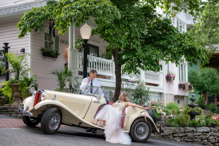 A bride and groom with a vintage roadster in front of the inn